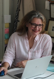 A smiling woman with shoulder-length wavy brown hair and glasses sits at a desk, wearing a light pink button-down shirt while operating a laptop and mouse.