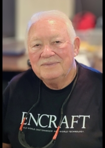 Close-up portrait of a man with white hair, a neat white mustache, wearing a dark T-shirt and smiling warmly.
