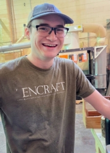 A smiling young man wearing a baseball cap, glasses, and a grey t-shirt with "ENCRAFT" printed across the front, standing in a workshop.