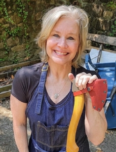 A smiling blonde woman wearing overalls and holding a carving axe. She is standing in front of an ivy-covered stone wall.
