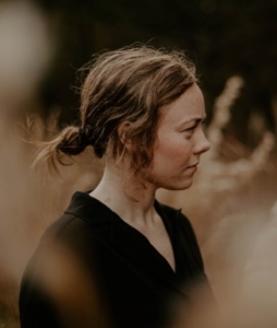 Side profile portrait of a woman with her brown hair tied back in a messy low ponytail, looking thoughtfully to the right against a soft-focus, outdoor background.