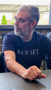 A man with short grey hair, a beard, and glasses sitting at a table in a dark t-shirt featuring the word "ENCRAFT" while looking off to the side.