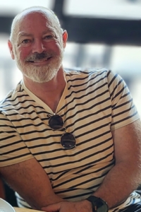 Close-up portrait of a smiling man with a short grey beard and shaved head, wearing a light-colored, dark-striped V-neck t-shirt with sunglasses hooked into the collar.