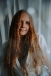 A woman with long, straight reddish-brown hair staring gently forward while wearing a white top, shot through a soft, hazy foreground element.